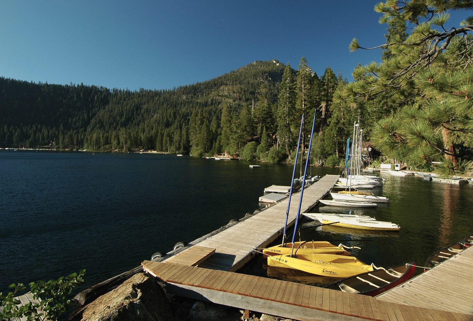 Small sailboats and yellow kayaks at a wooden boat dock on a blue mountain lake, pine-covered slopes behind