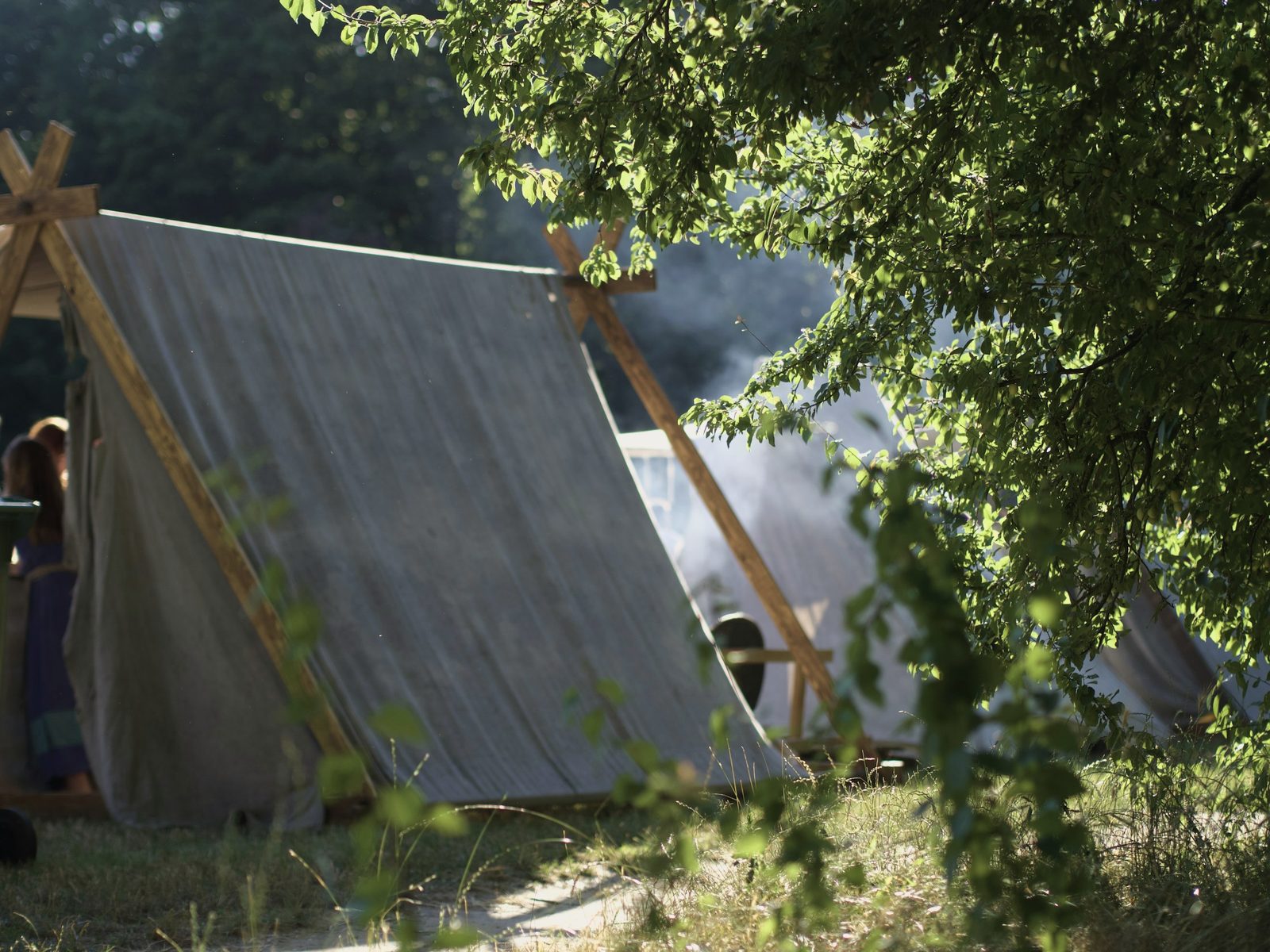 Canvas A-frame tents on wooden poles in a sunlit wooded campsite, a haze of campfire smoke behind